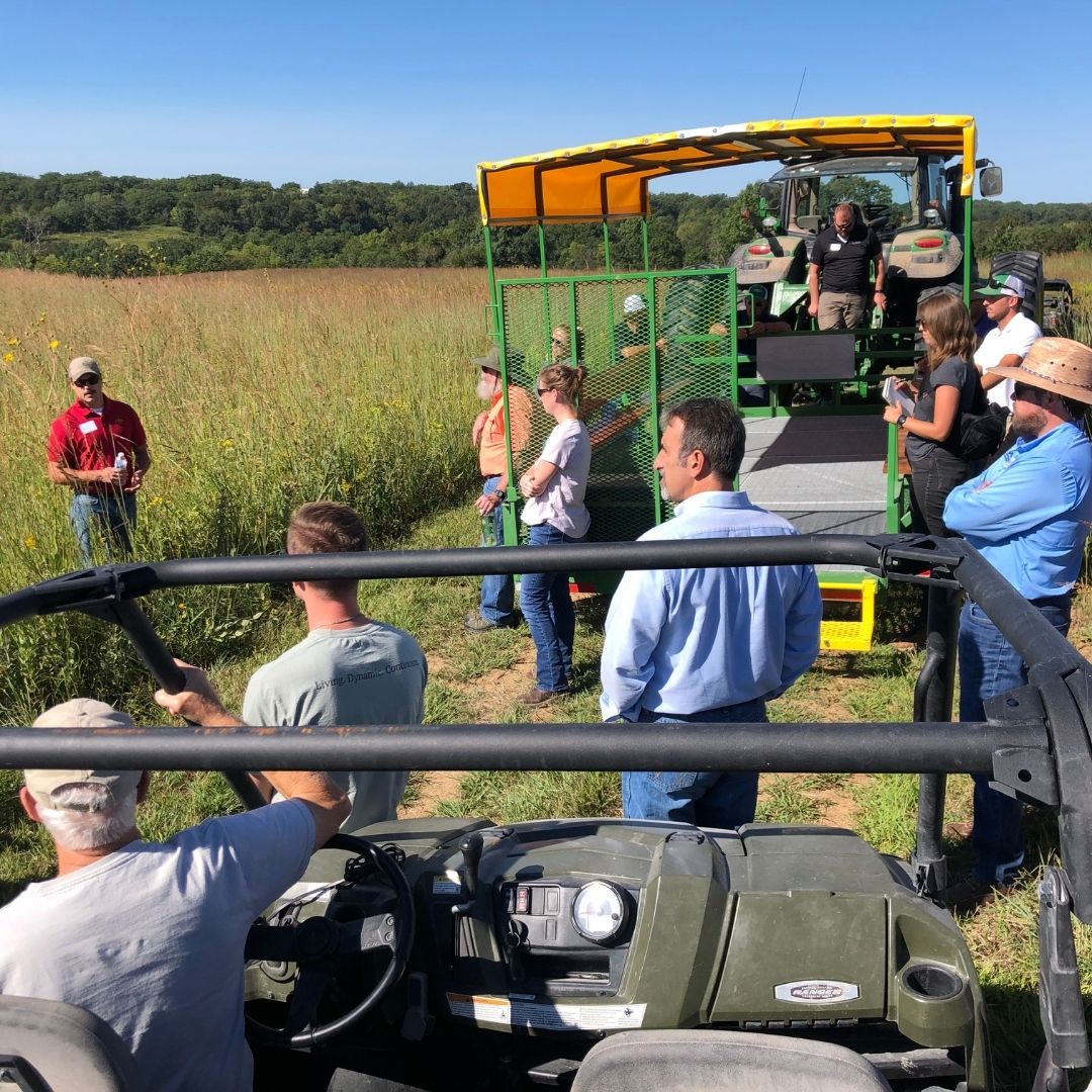 Photo of man in a prairie speaking to group of field trip attendees, viewing from ATVs and trailers. 